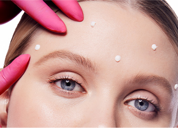 A pink medical gloved hand rubbing in Dynamic Wrinkle Limiter topical cream to targeted spots on a blonde woman’s forehead